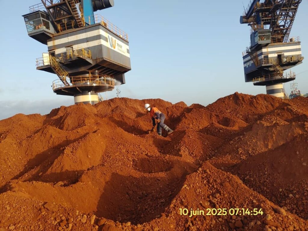 Worker amidst large piles of reddish-brown ore under industrial cranes.