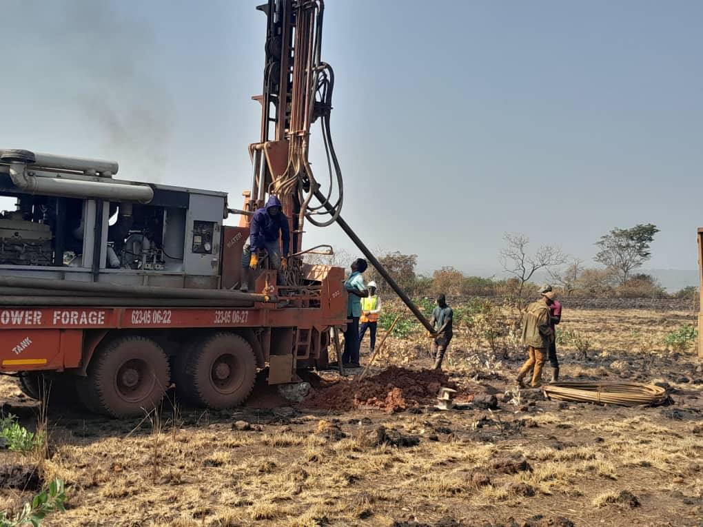 Workers operate a drilling rig in a dry, rural field under clear skies.